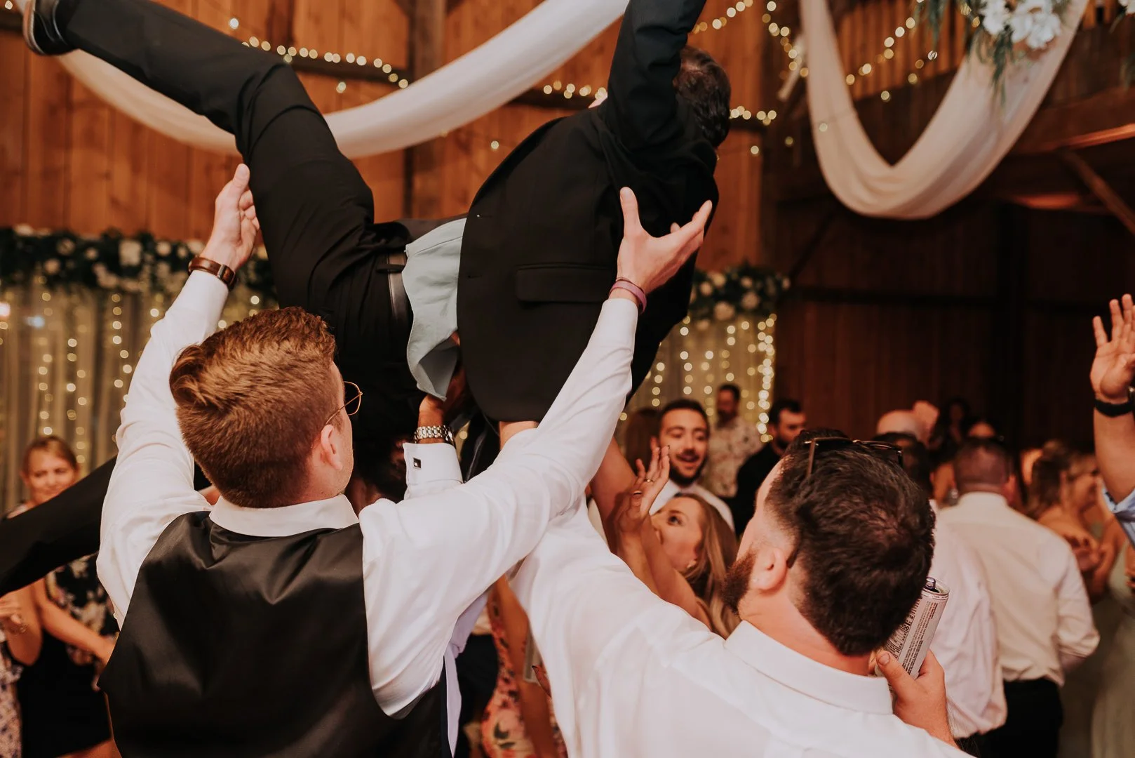 Group of people celebrating and lifting a man in a black suit at a party or wedding reception with warm lighting and decorations.
