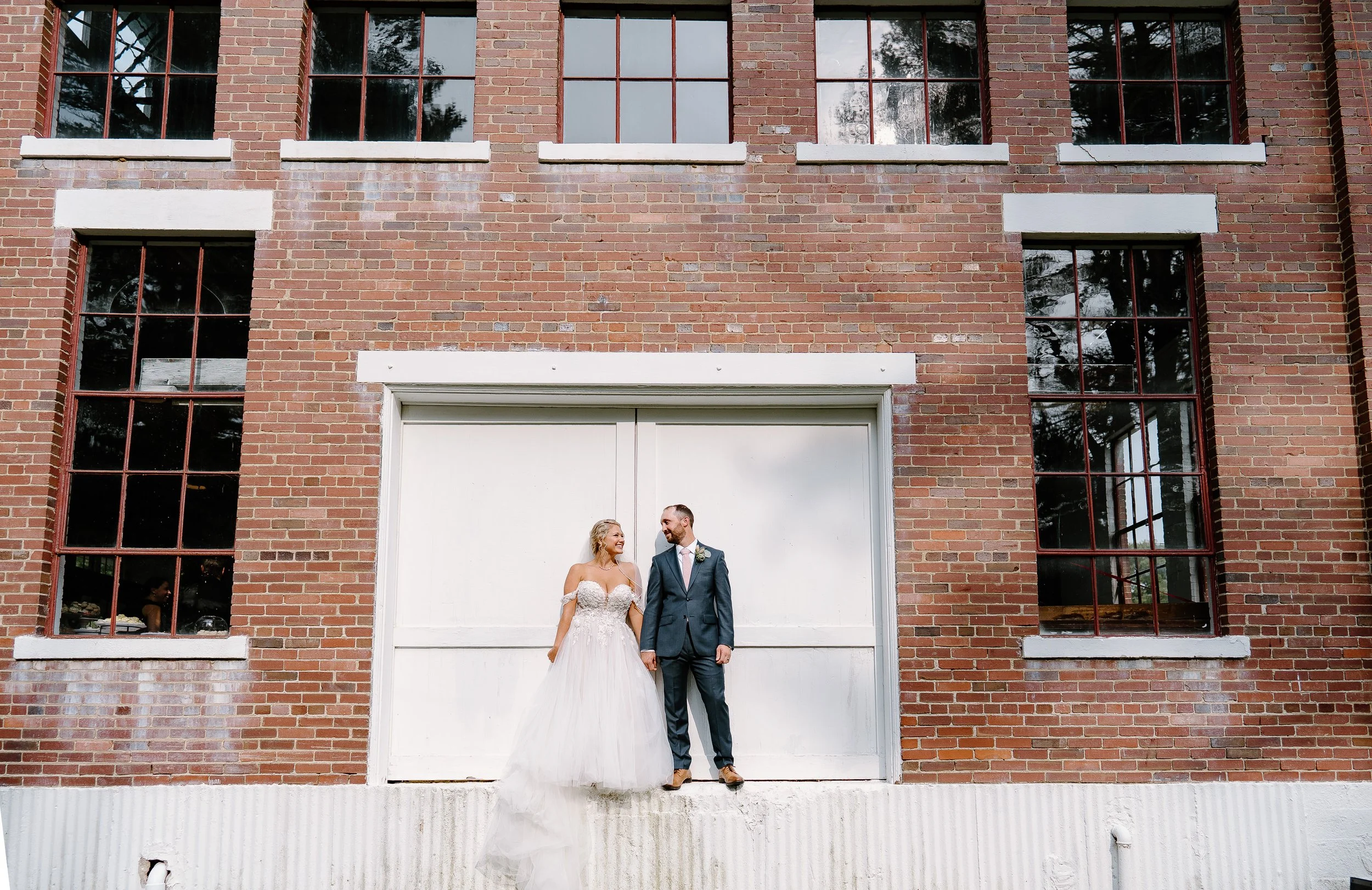 A bride and groom holding hands, standing in front of a large white garage door on a brick building with tall windows, outdoors on a sunny day.