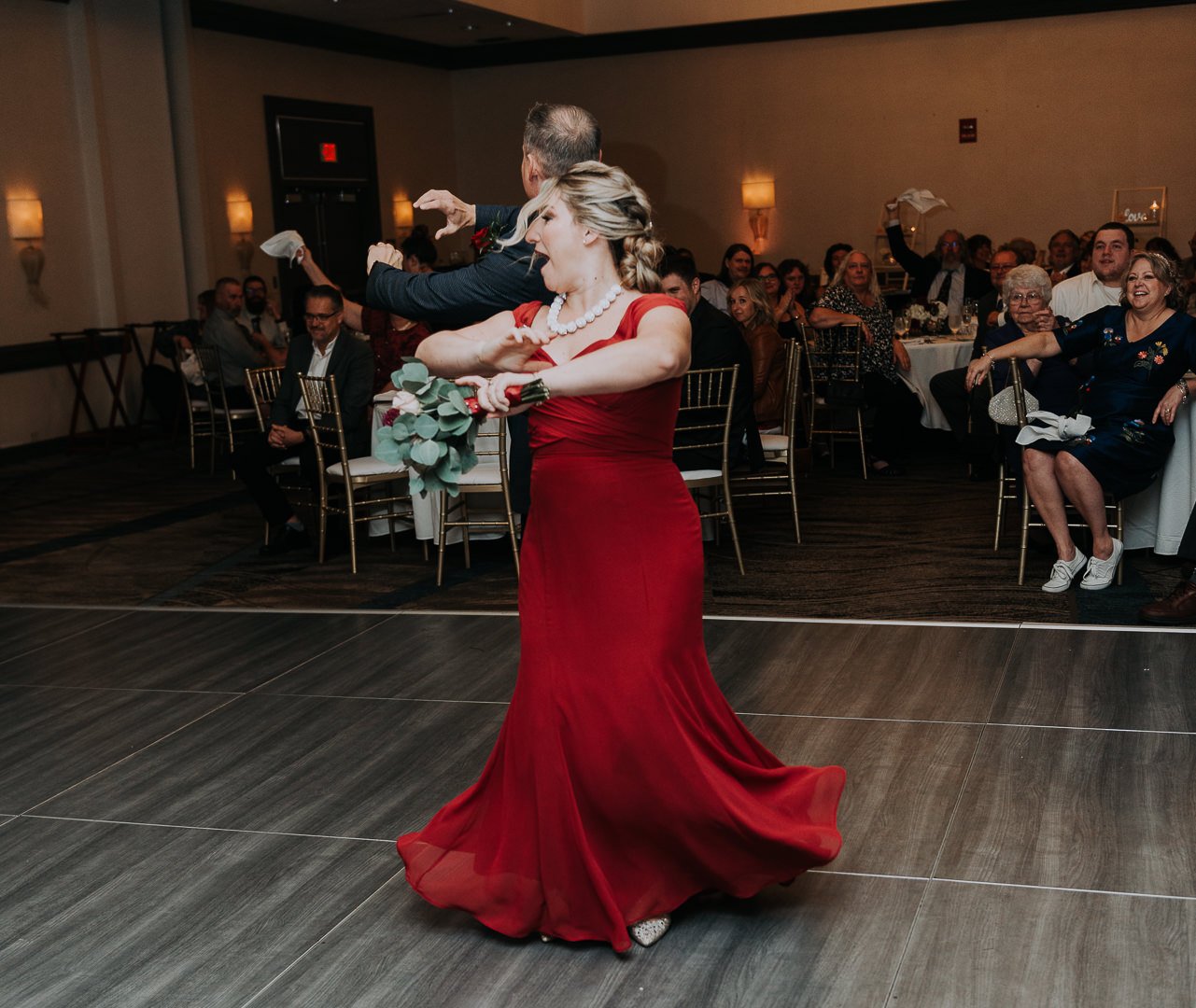 A woman in a red evening gown dancing with a man in a tuxedo at a wedding reception, with guests seated at tables in the background.
