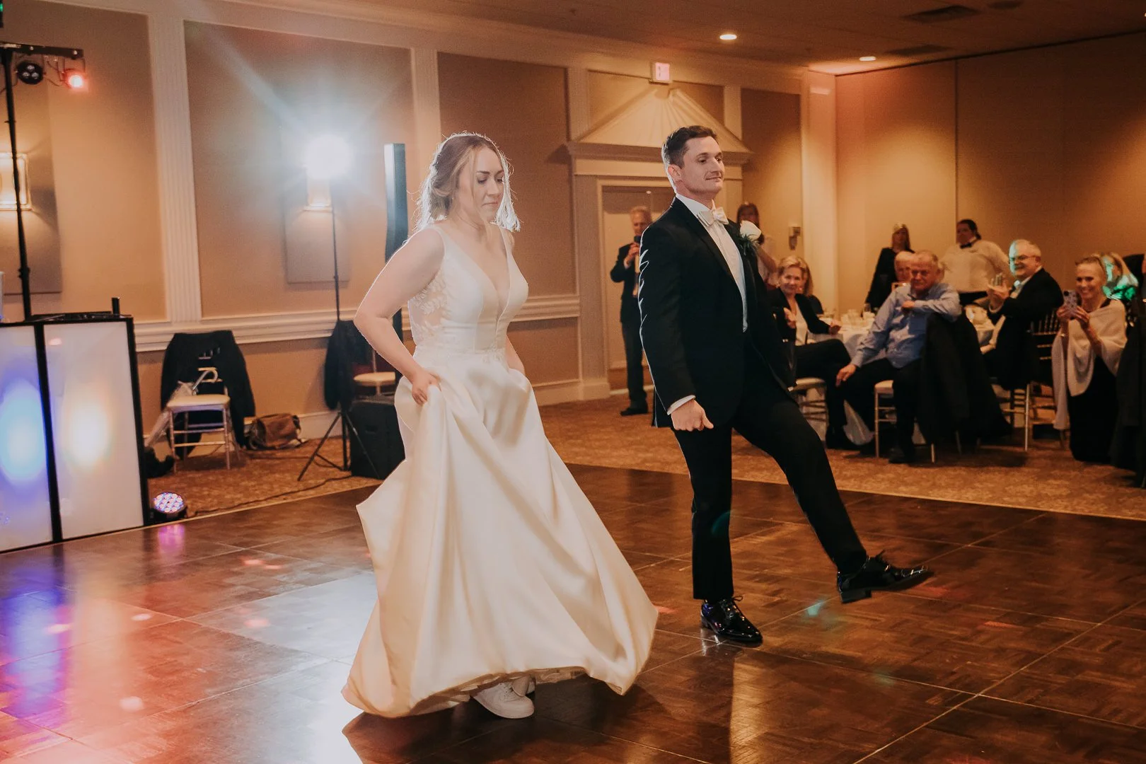A bride and groom are dancing at their wedding reception in a banquet hall, surrounded by seated guests watching and smiling.