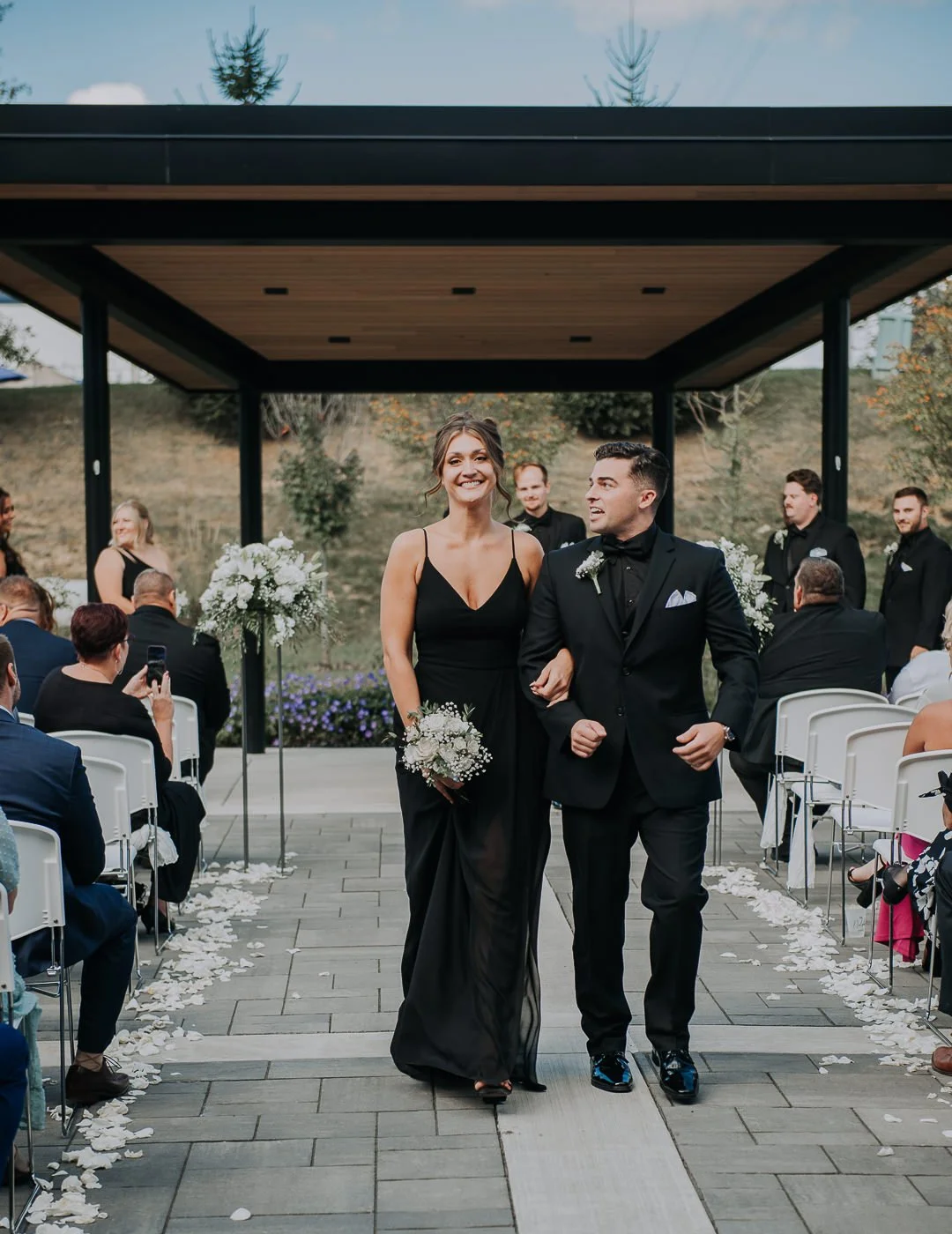 A bride and groom walk down the aisle at their outdoor wedding, smiling, with guests seated on either side. The bride wears a black gown and the groom a black tuxedo. Guests are taking photos, and there are flower arrangements and petals along the ai