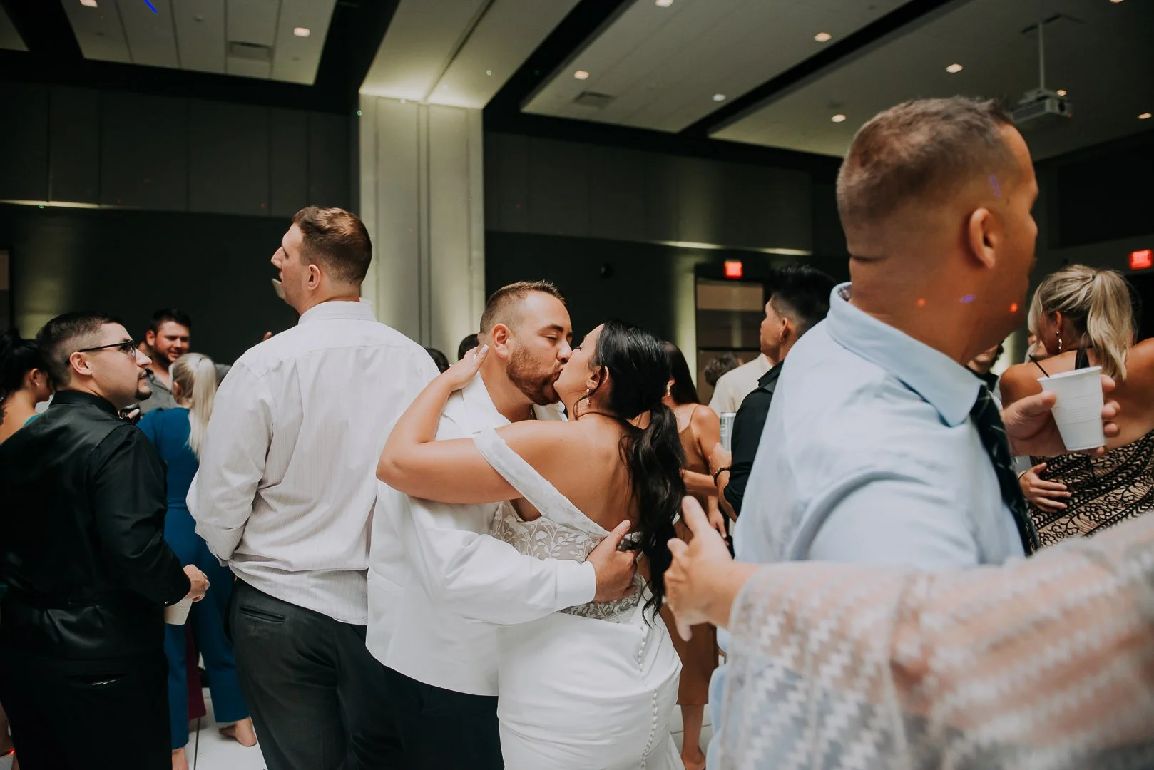 A couple sharing a kiss during a celebration or party, with other people gathered around in a large indoor venue.