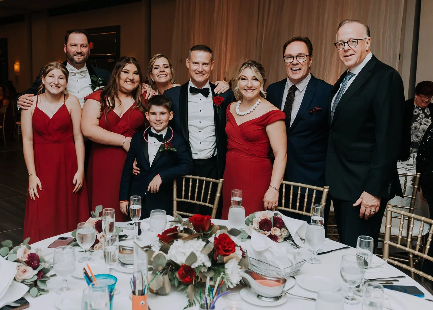 A group of ten people, including men, women, and a boy, dressed in formal attire, posing together at a celebration or wedding reception. They are smiling and standing behind a decorated table with floral arrangements and glassware.