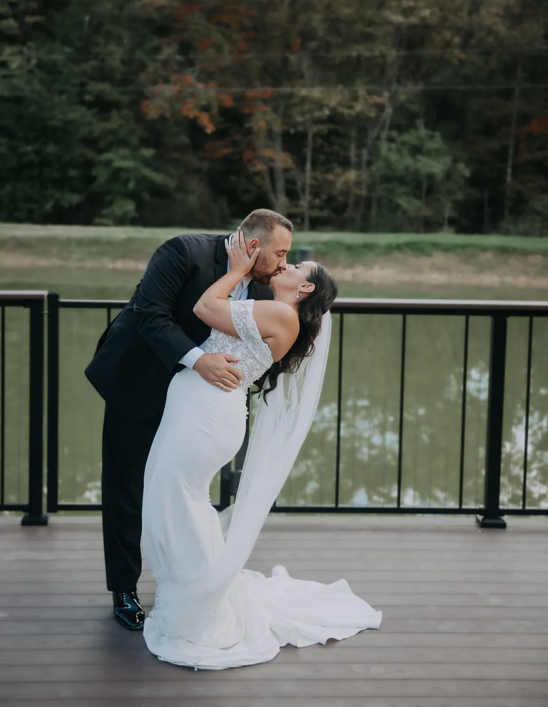A newlywed couple sharing a kiss on a wooden deck beside a lake with trees in the background.