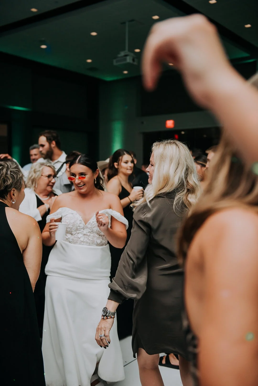 People dancing and socializing at a party or celebration, with a woman in a white dress and red sunglasses in focus.