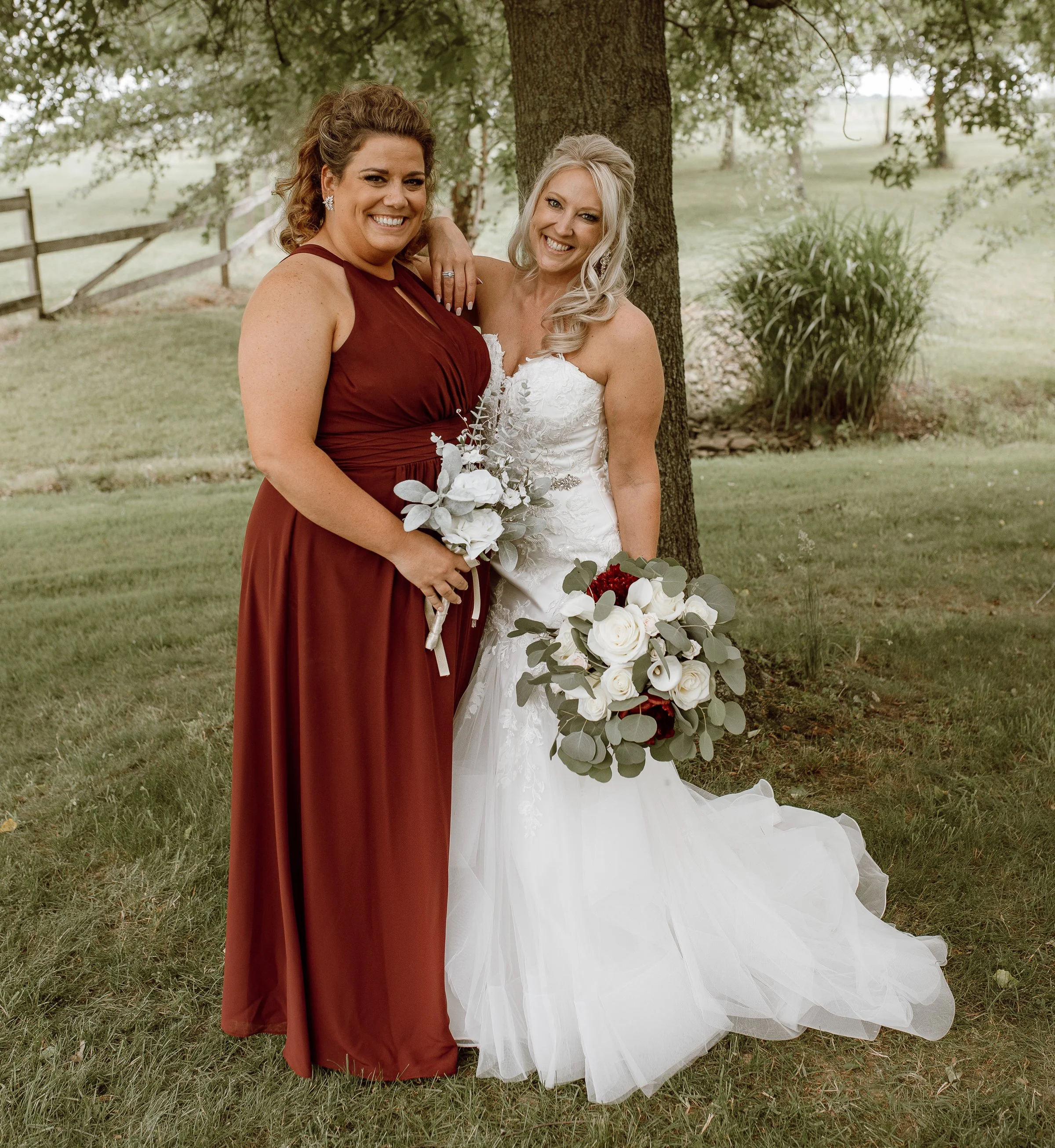 Two women, one in a white bridal gown and the other in a burgundy dress, standing outdoors near a tree, holding bouquets of flowers, smiling, with a grassy landscape and trees in the background.