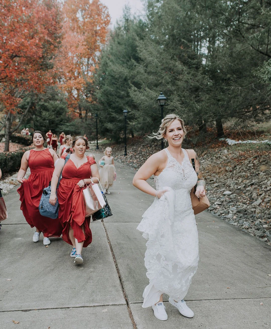Group of women, some in red dresses and one in a white wedding dress, walking outdoors on a paved pathway surrounded by autumn trees and greenery during the daytime.