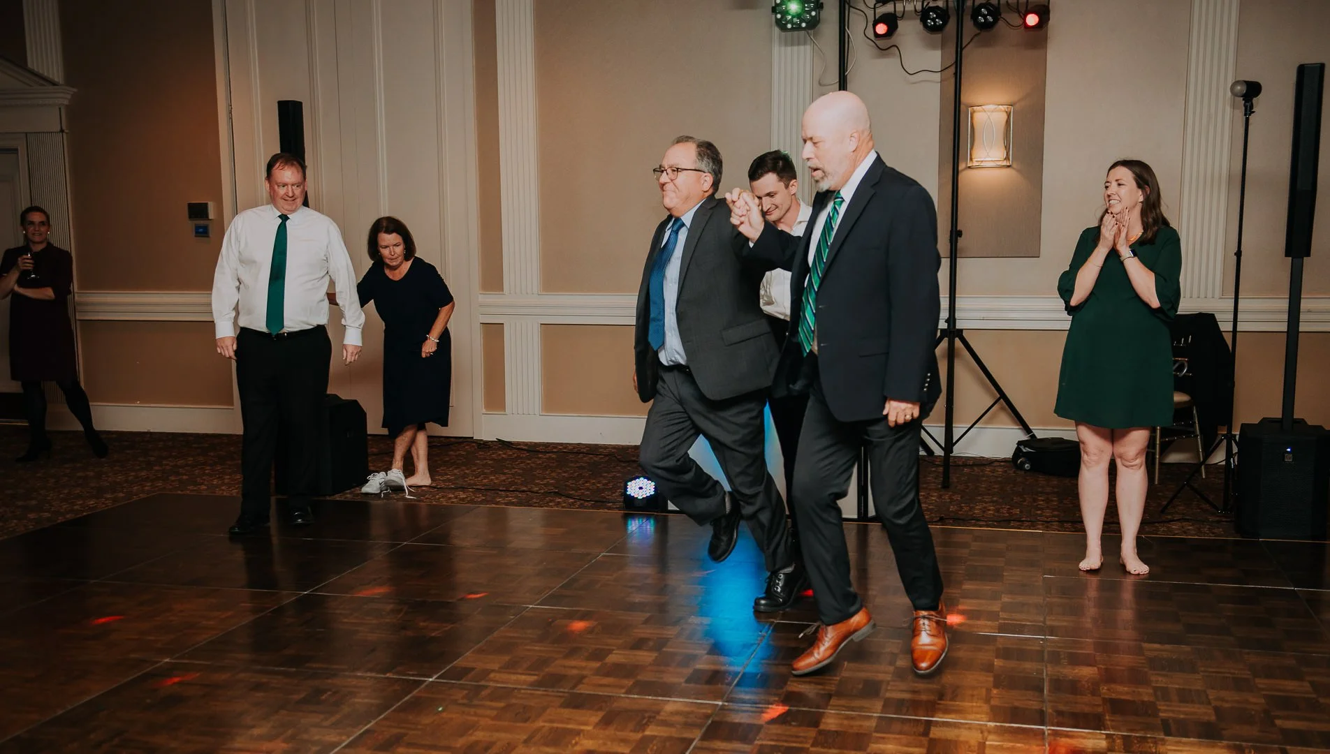 Group of adults dancing and celebrating at an event in a hotel ballroom with a dance floor, some people are smiling and clapping, mood is joyful and festive.