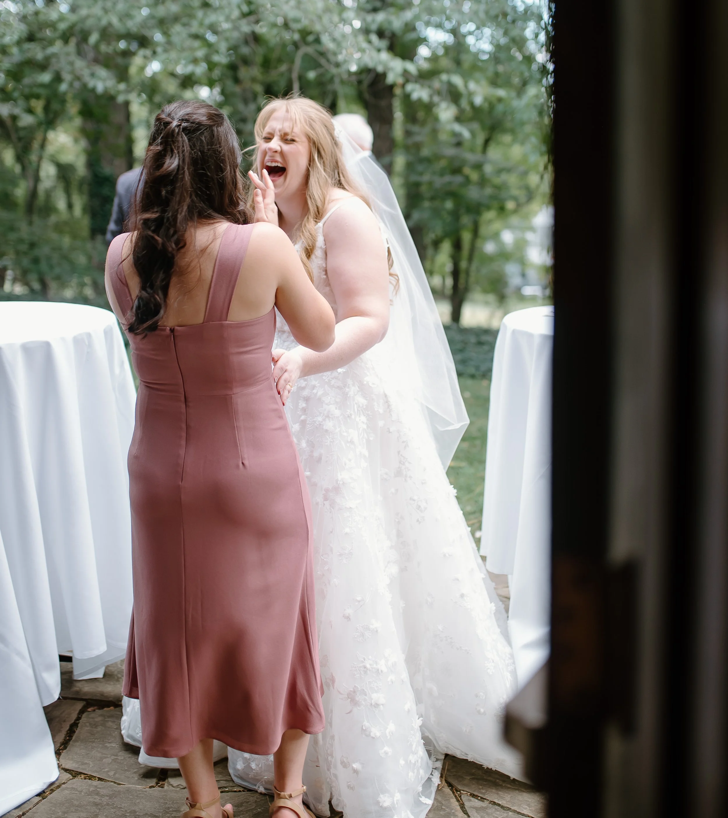Two women, one wearing a pink dress and the other a white wedding gown, appear to be emotional and crying during a wedding ceremony outdoors surrounded by trees.