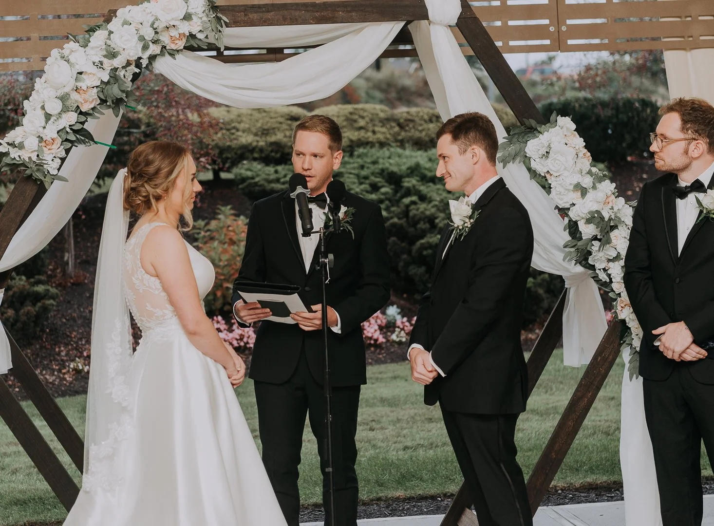 A wedding ceremony outdoors with a bride and groom standing under a wooden arch decorated with white and blush flowers and draped white fabric, officiant with a tablet, and two groomsmen in black tuxedos.