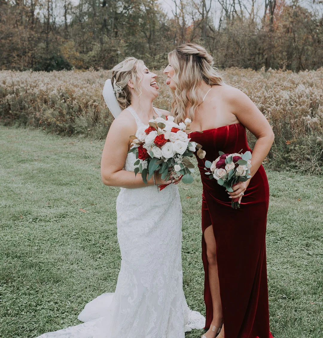 Two women, one in a white wedding dress and the other in a red velvet dress, laugh together outdoors, holding bouquets, in a grassy field with trees in the background.