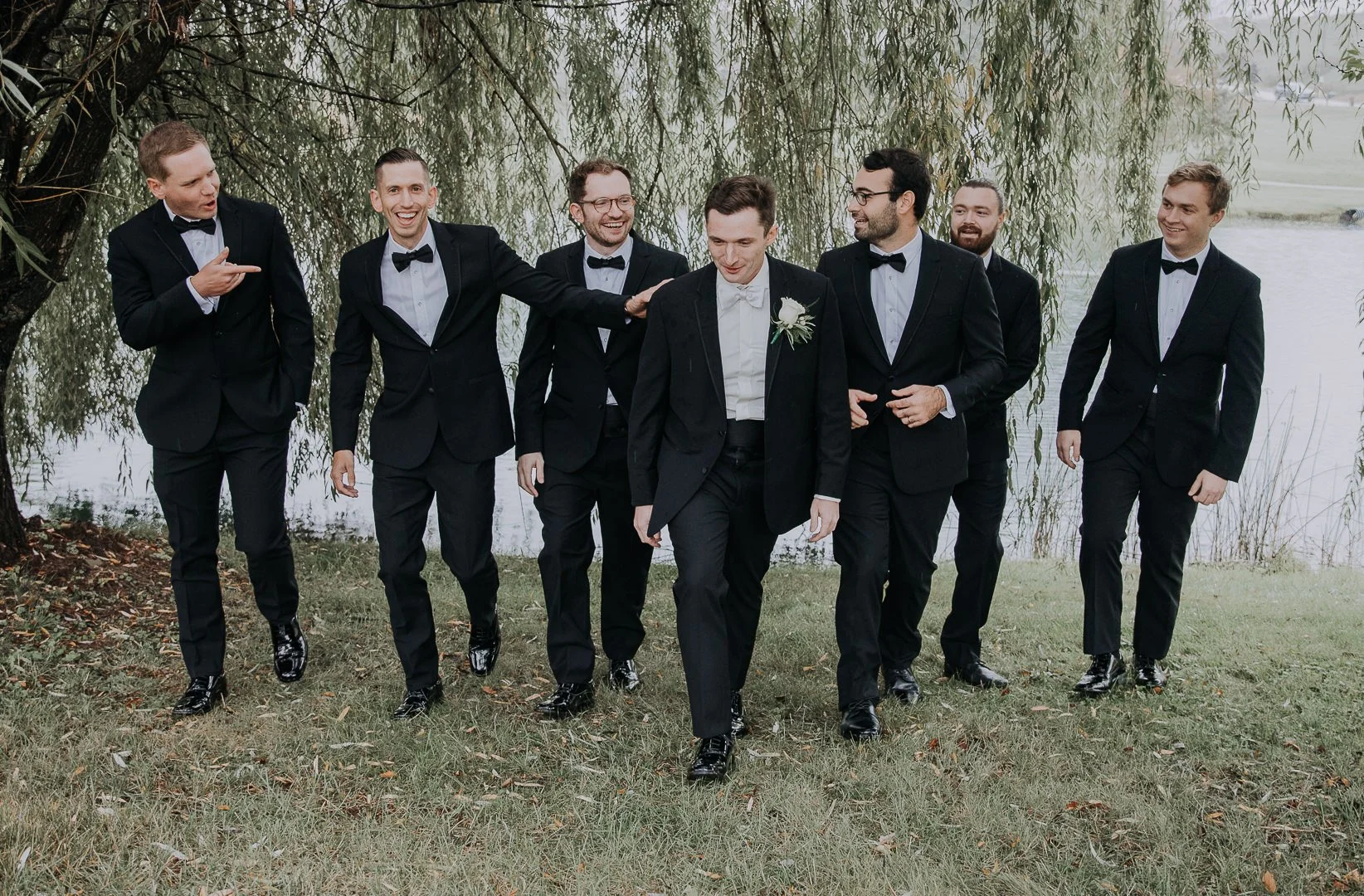 A group of eight men in tuxedos walking together outdoors near a lake, with one man wearing a boutonniere, under a large tree with hanging branches.