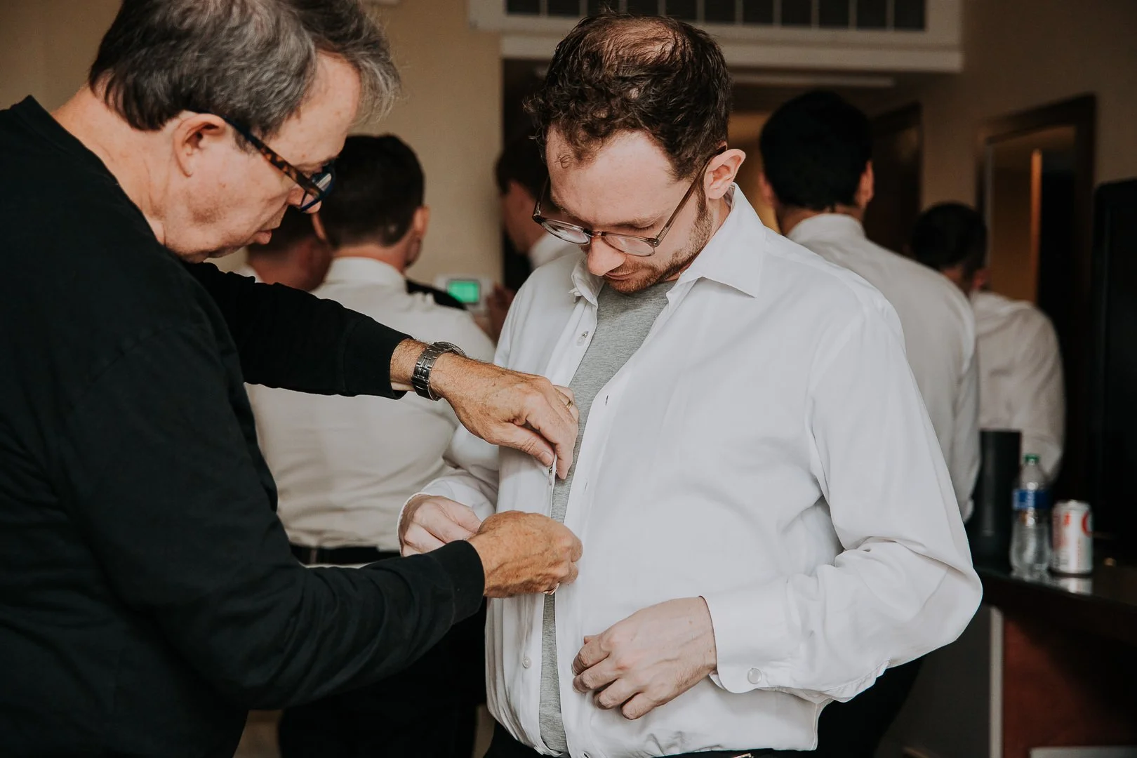 Two men, one assisting the other with a white dress shirt at an indoor gathering, with several people in white shirts in the background.