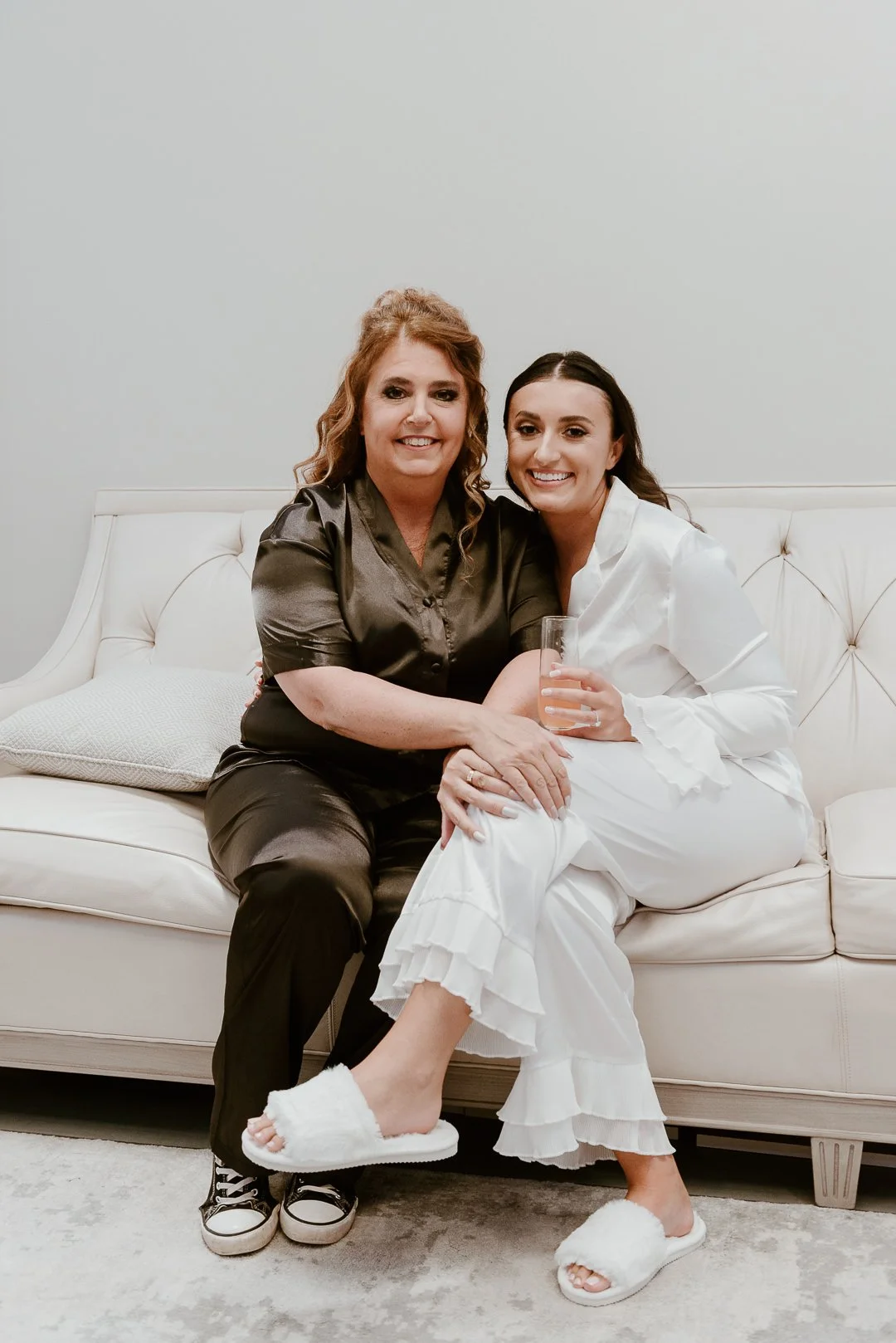 Two women sitting on a white couch, smiling and embracing each other. One woman is wearing a black satin pajama set and black sneakers, the other is in white pajamas and fluffy slippers, holding a glass of pink drink.