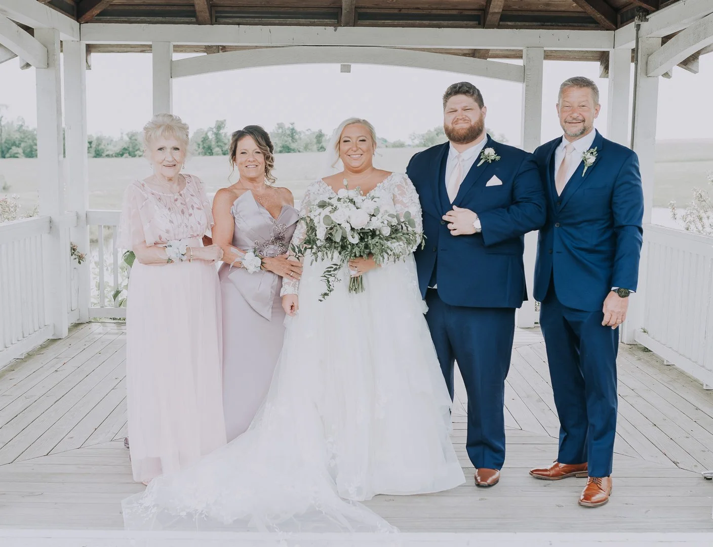 A bride in a white wedding gown holding a bouquet, flanked by a woman in a lavender dress, a woman in a pink dress, and two men in blue suits at an outdoor wedding ceremony