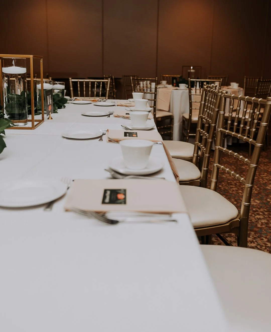 A banquet table set with white plates, cups, silverware, and beige napkins, with gold Chiavari chairs around it, in a formal dining room.