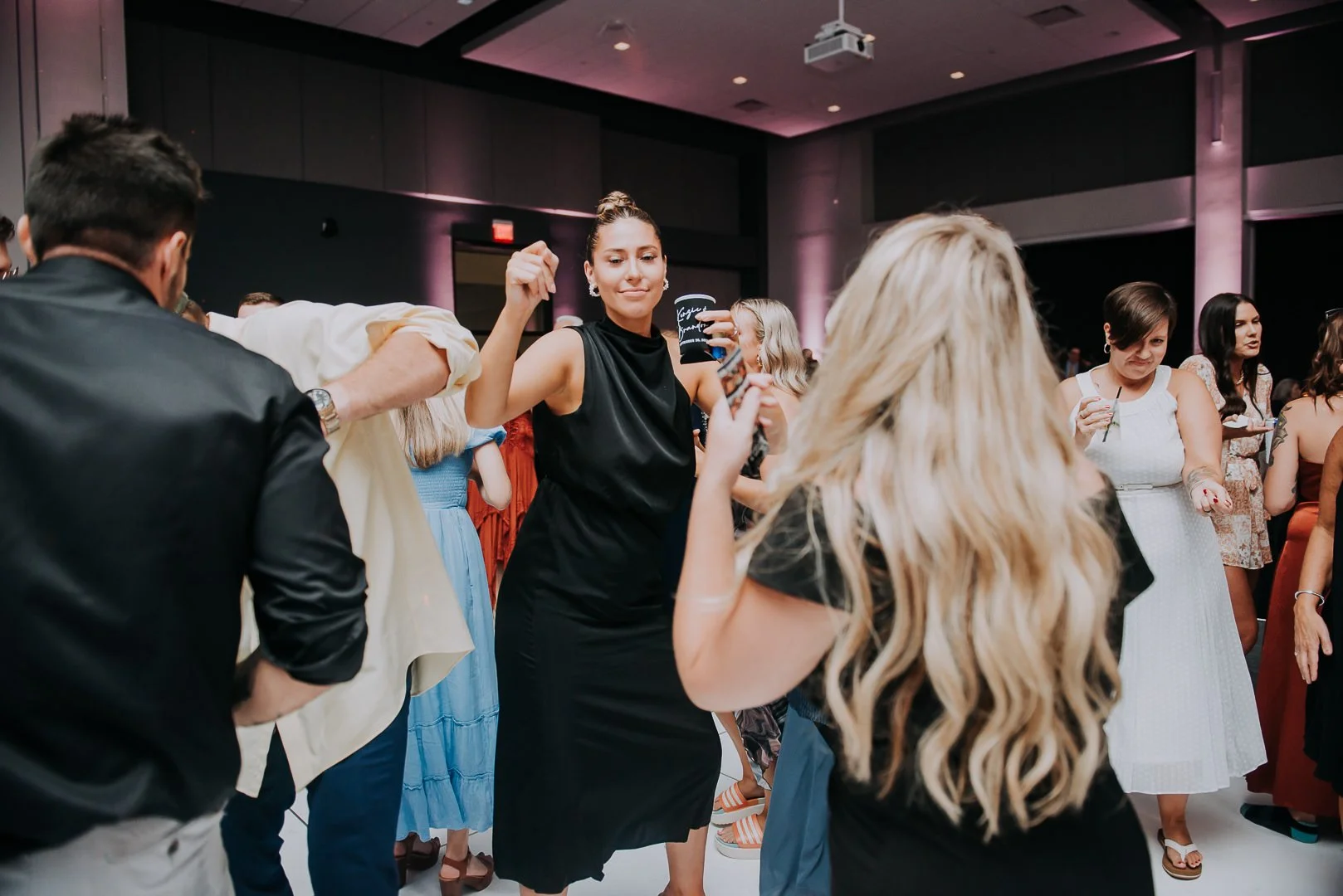 People dancing and socializing at a party or event in a large indoor hall with dark walls and purple lighting.