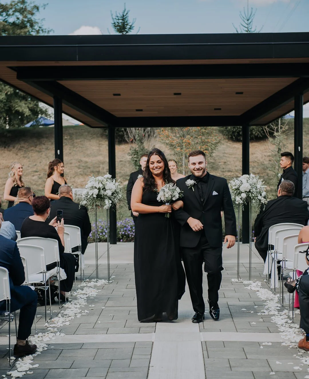 A happy couple walks down the aisle at their wedding ceremony, surrounded by seated guests and floral arrangements, outdoors under a modern pavilion with trees in the background.