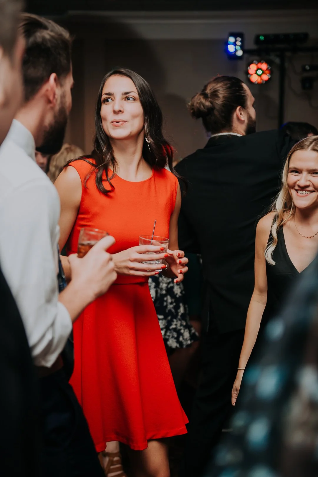 People socializing at a party, two women in black and red dresses chatting, holding drinks, smiling, with a DJ or music setup in the background.