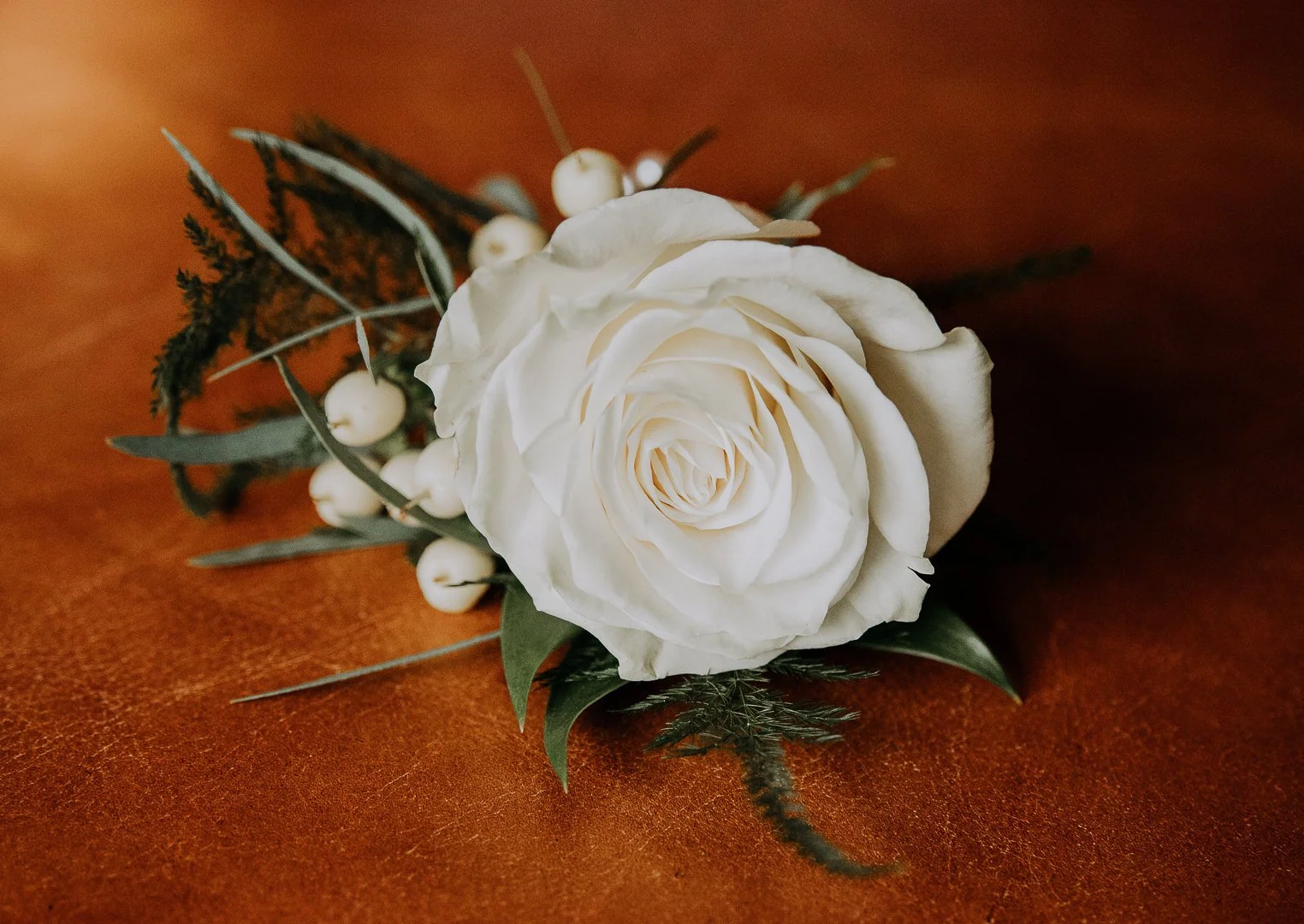 A white flower boutonniere with green leaves, white berries, and fern-like greenery on a brown surface.