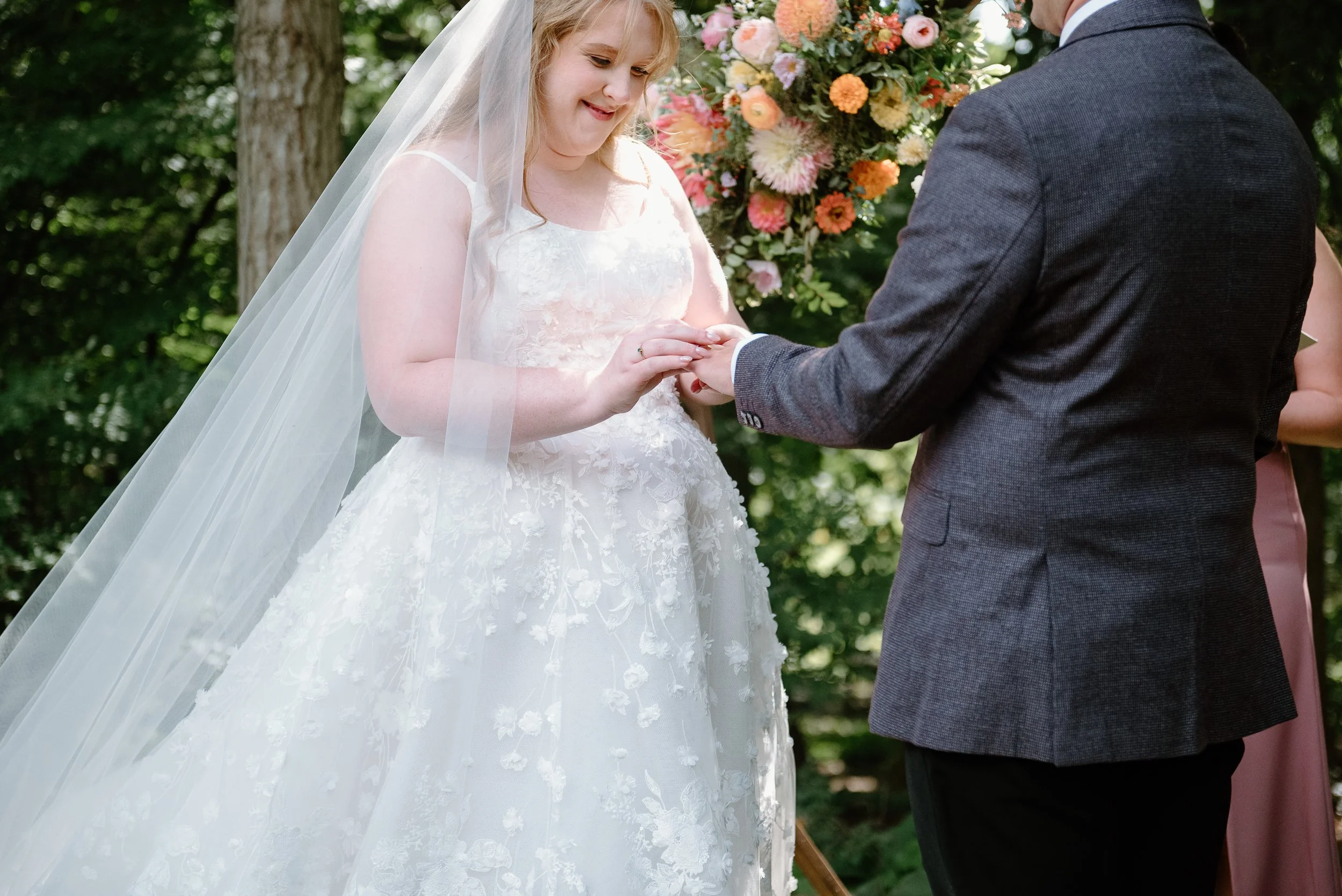 A bride in a white wedding dress and veil exchanges rings with a groom wearing a dark gray suit during an outdoor wedding ceremony, with a colorful flower arrangement in the background.