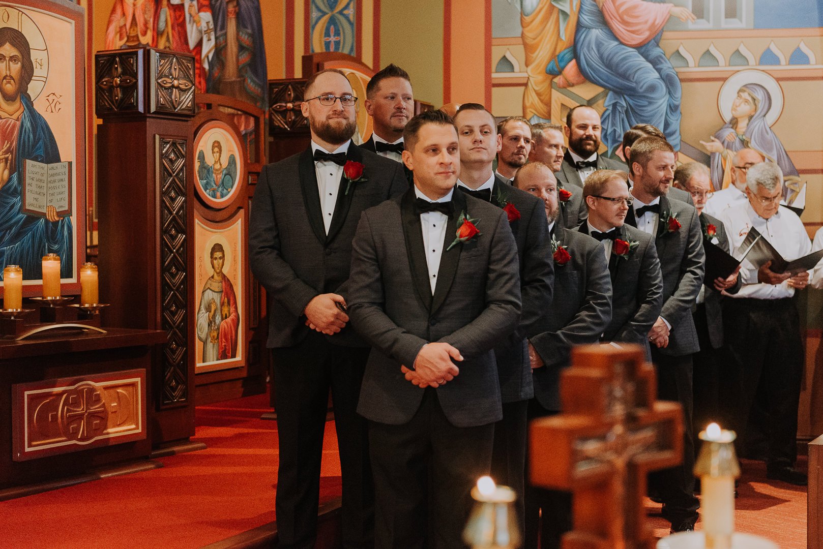 A group of men wearing suits and bow ties, standing inside an Orthodox church with religious icons and candles on the altar, during a wedding ceremony.