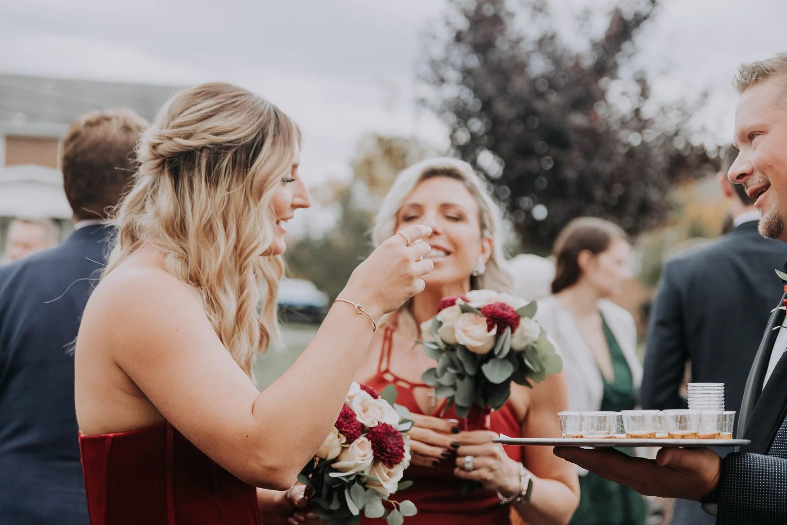 A woman in a red dress feeds a woman holding a bouquet of flowers at an outdoor wedding reception.