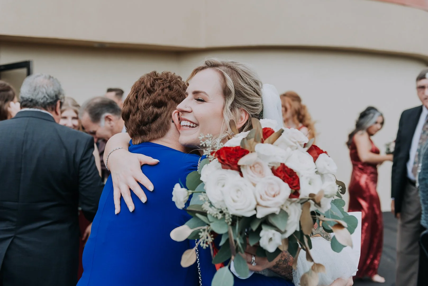 A woman in a wedding dress holding a bouquet of white and red roses, hugging a woman in a blue dress, at a wedding reception or celebration with guests in the background.