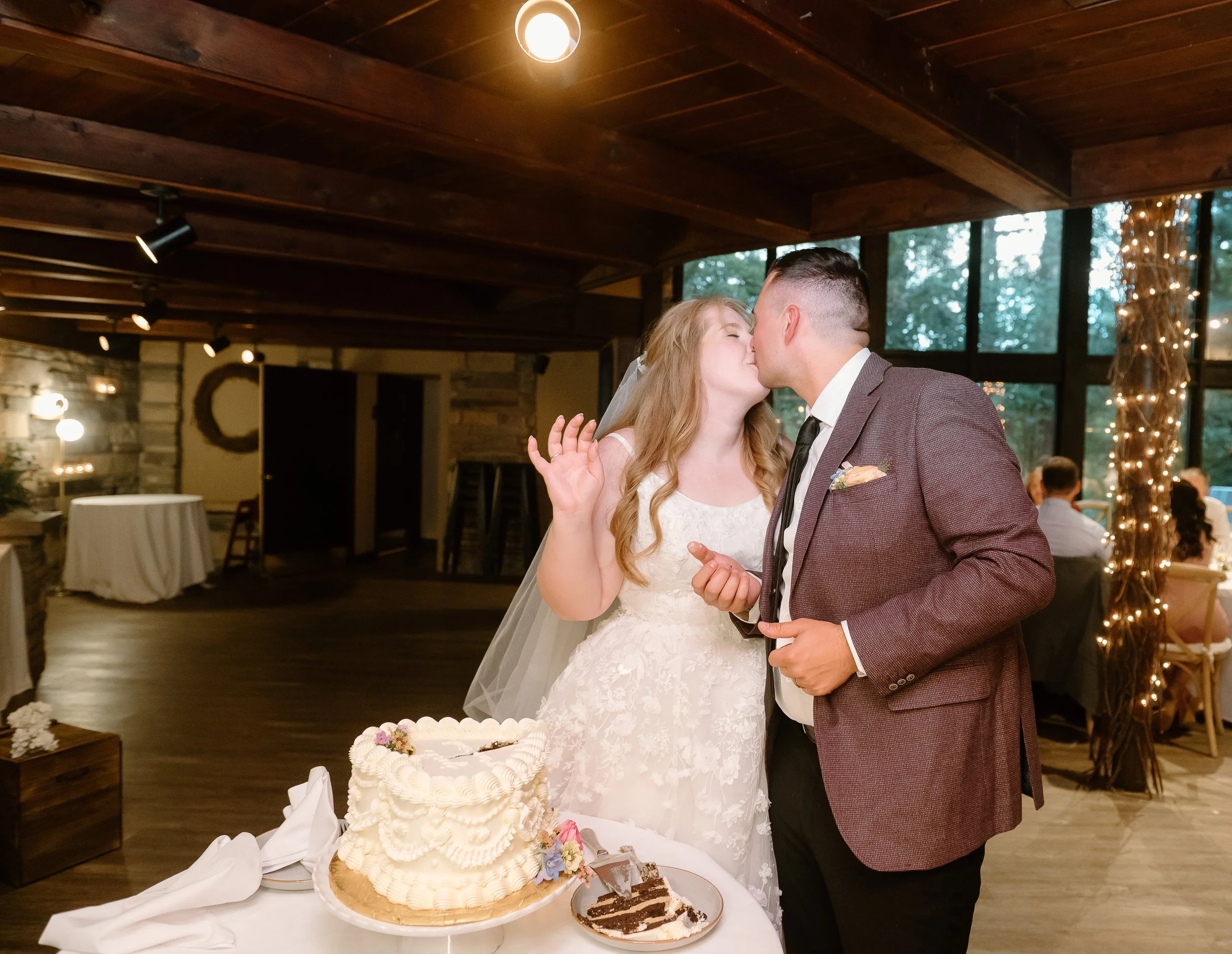A bride and groom share a kiss at their wedding reception, with a wedding cake on the table in front of them, in a warmly decorated indoor venue with string lights and large windows.