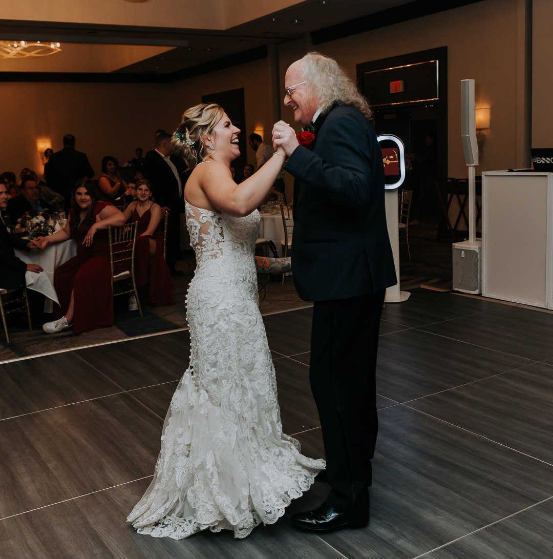 A bride and an older man dance together at a wedding reception, holding hands and smiling, with guests seated at tables in the background.