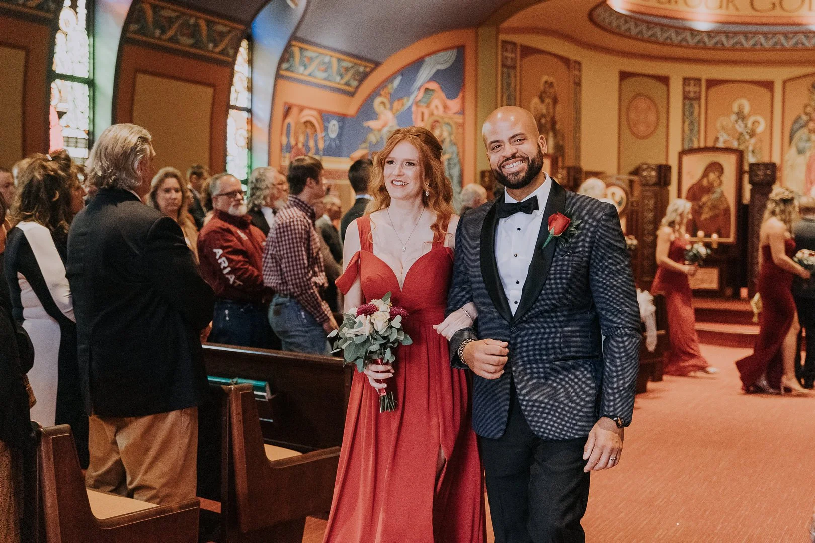 A smiling bride with long red hair in a red gown holding a bouquet, standing next to a smiling groom in a dark tuxedo with a boutonniere, inside a church during a wedding ceremony with guests in the background.