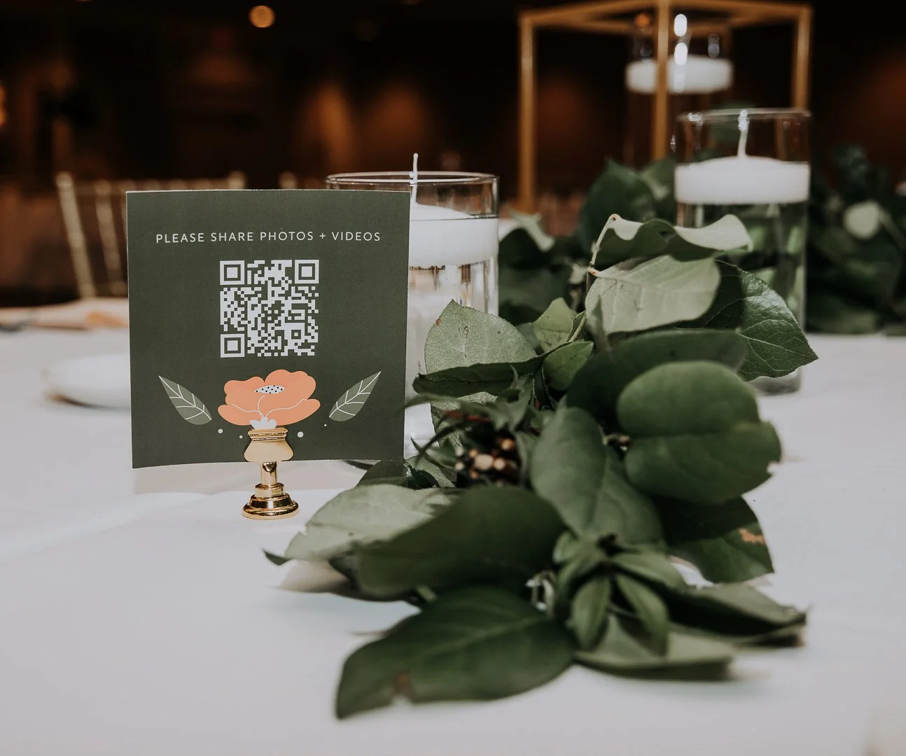 Table centerpiece with greenery, water candles, and a sign with a QR code for sharing photos and videos at an event.