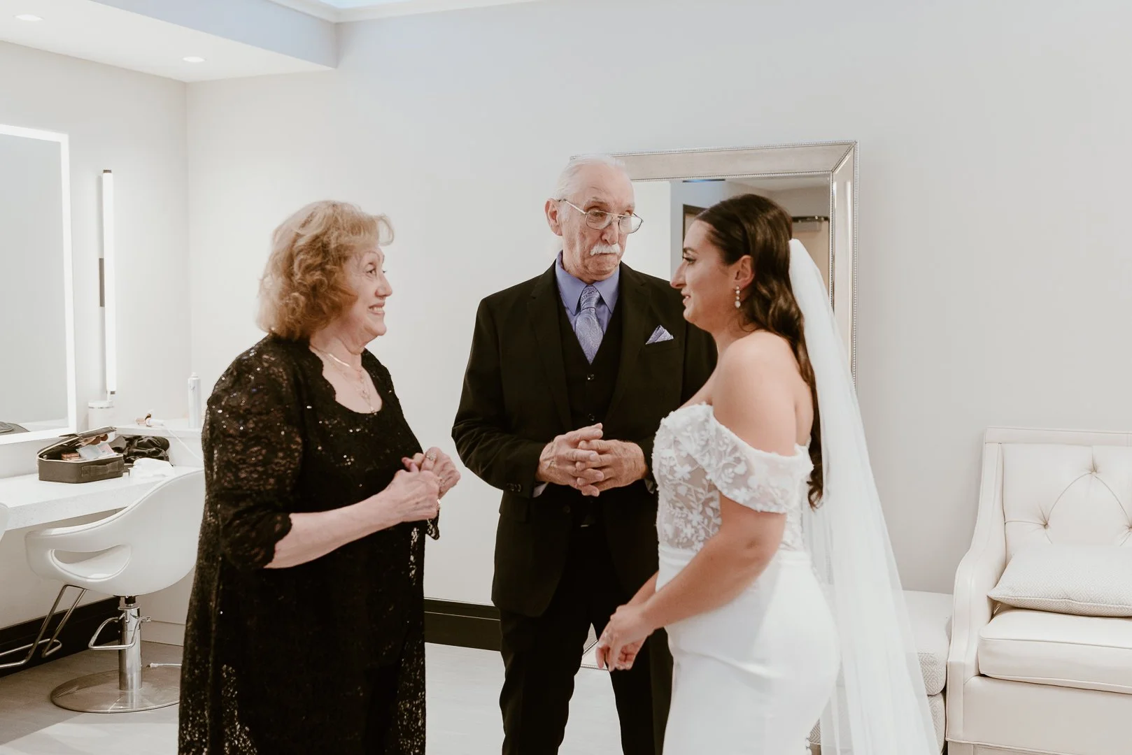 A bride in a white off-the-shoulder wedding dress with a veil, talking to an older woman in a black dress, and a man in a black suit with glasses, in a room with white walls and a mirror.