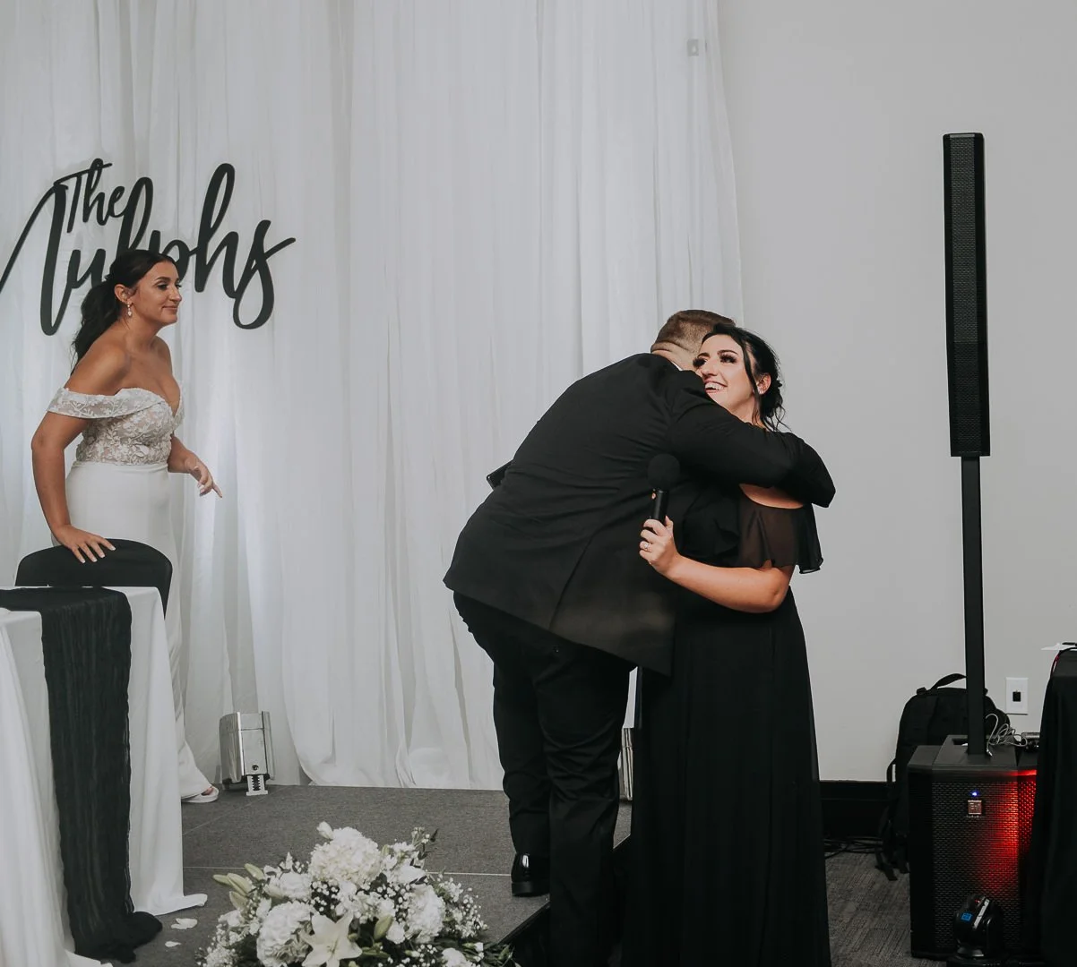 A woman receiving a hug from a man holding a microphone during a wedding reception, with another woman standing nearby near a decorated table and flowers, and a white curtain backdrop with the words "The Tugocks".