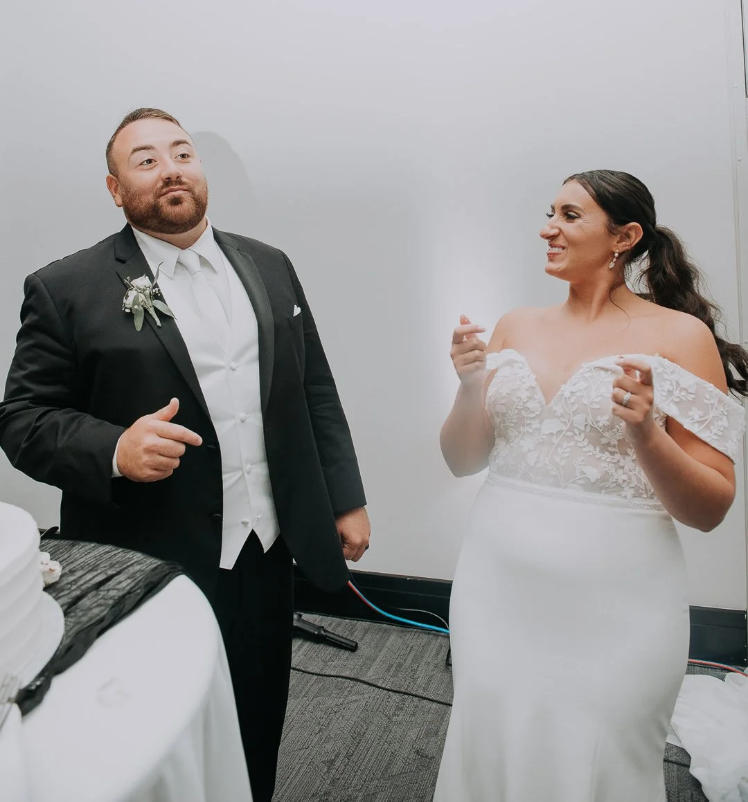 A bride and groom stand in a room, with the groom dressed in a black tuxedo and the bride in a white lace wedding gown, both smiling and looking at each other.