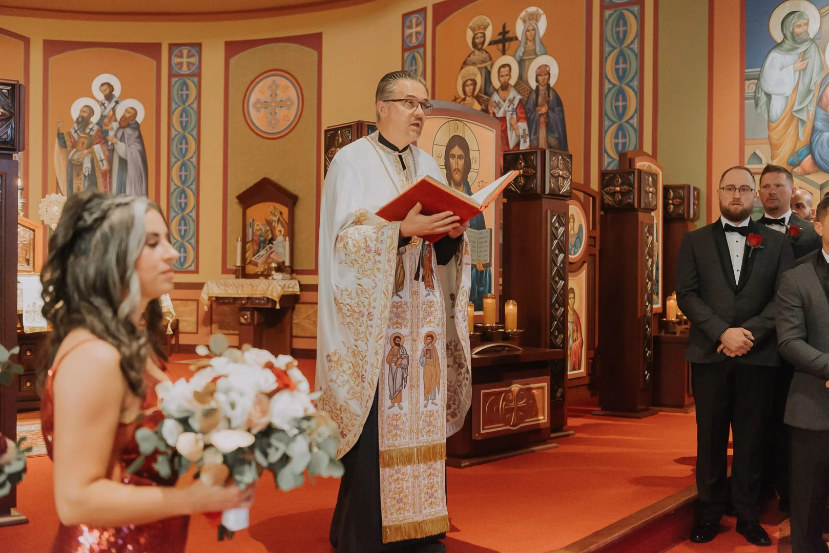 A wedding ceremony inside a Christian church. The bride, with wavy brown hair and wearing a red dress, is holding a bouquet of white and red flowers. The priest, dressed in ornate white and gold vestments, is standing and reading from a red book. Sev