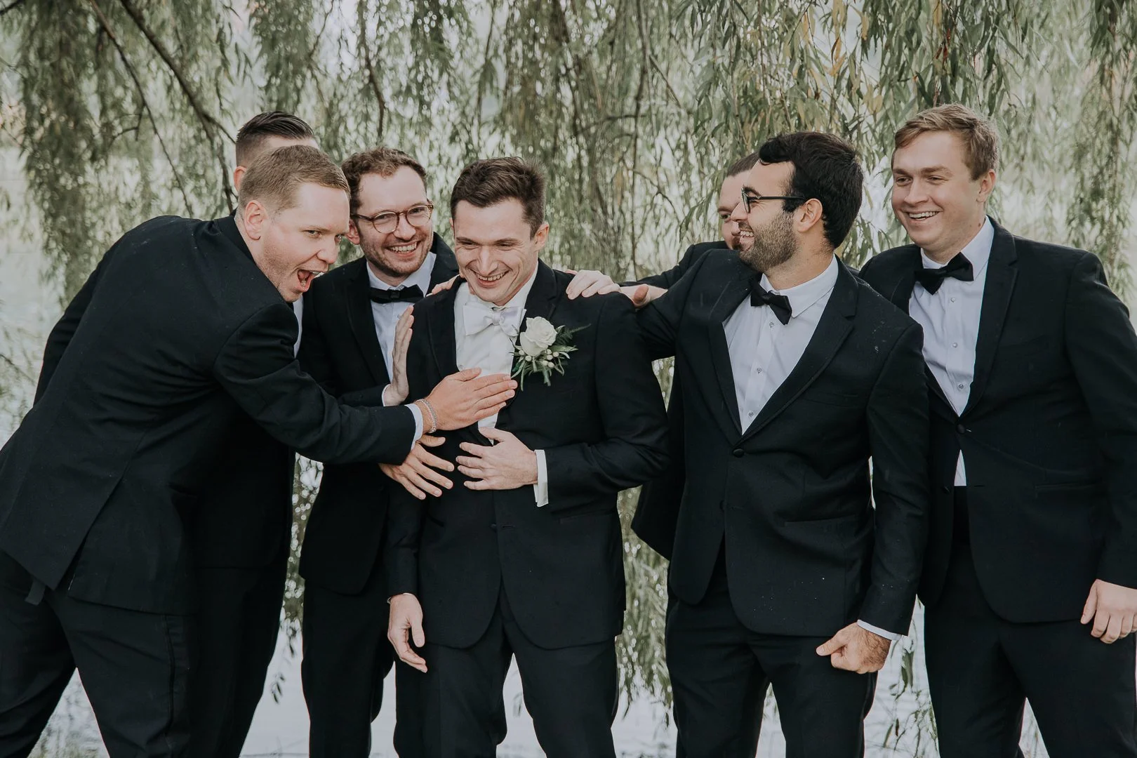 Group of men in tuxedos at a wedding, celebrating and smiling outdoors.