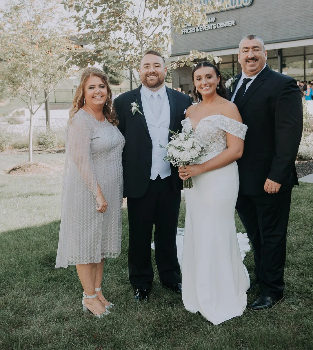 Group of four people standing outdoors on a grassy area, dressed in formal attire, with a bride in a white wedding gown holding a bouquet, and a groom in a black tuxedo. There are trees and a building with the sign 'Offices & Events Center' in the ba