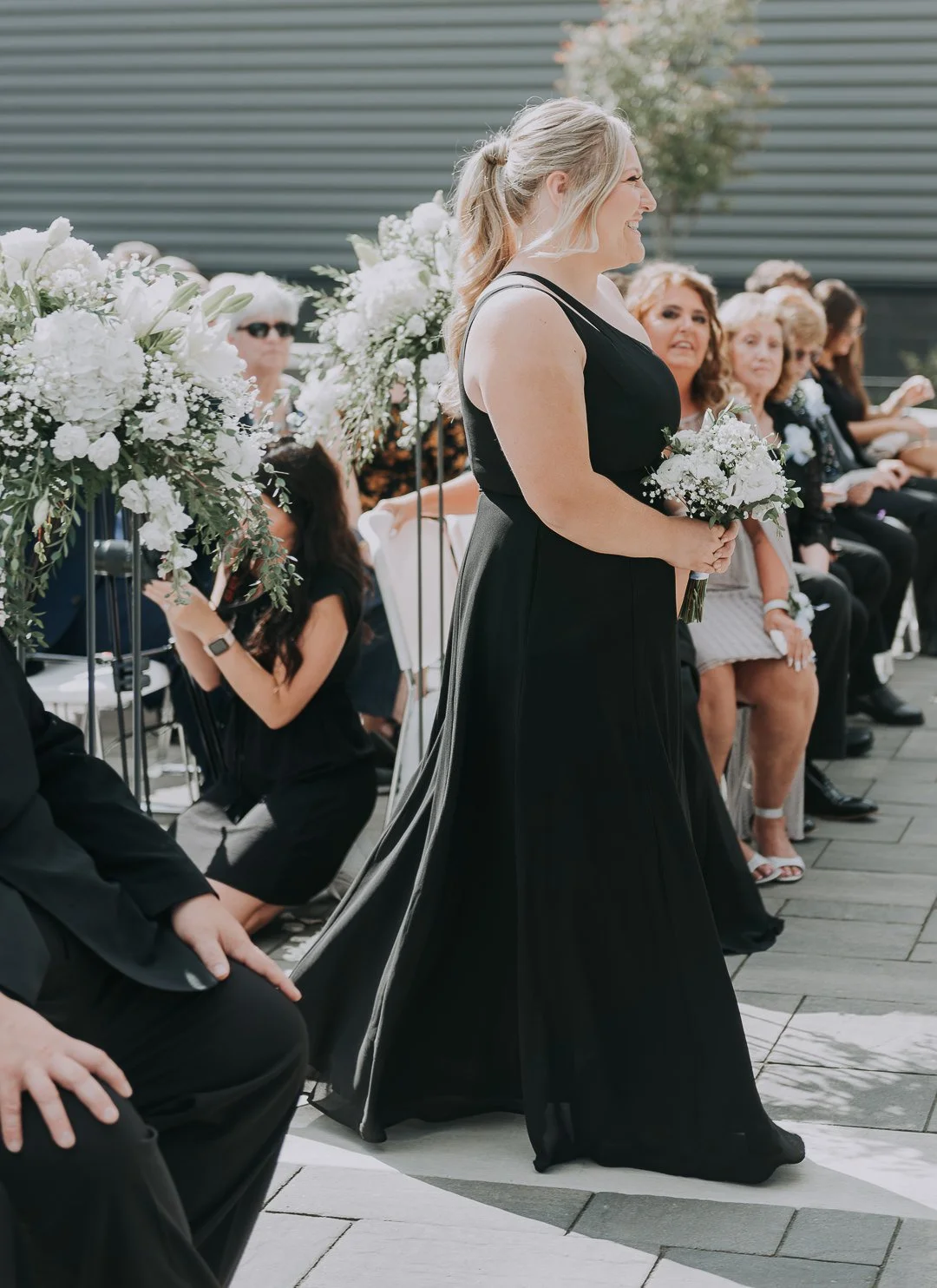A woman in a black dress holding a bouquet of white flowers at an outdoor wedding ceremony.