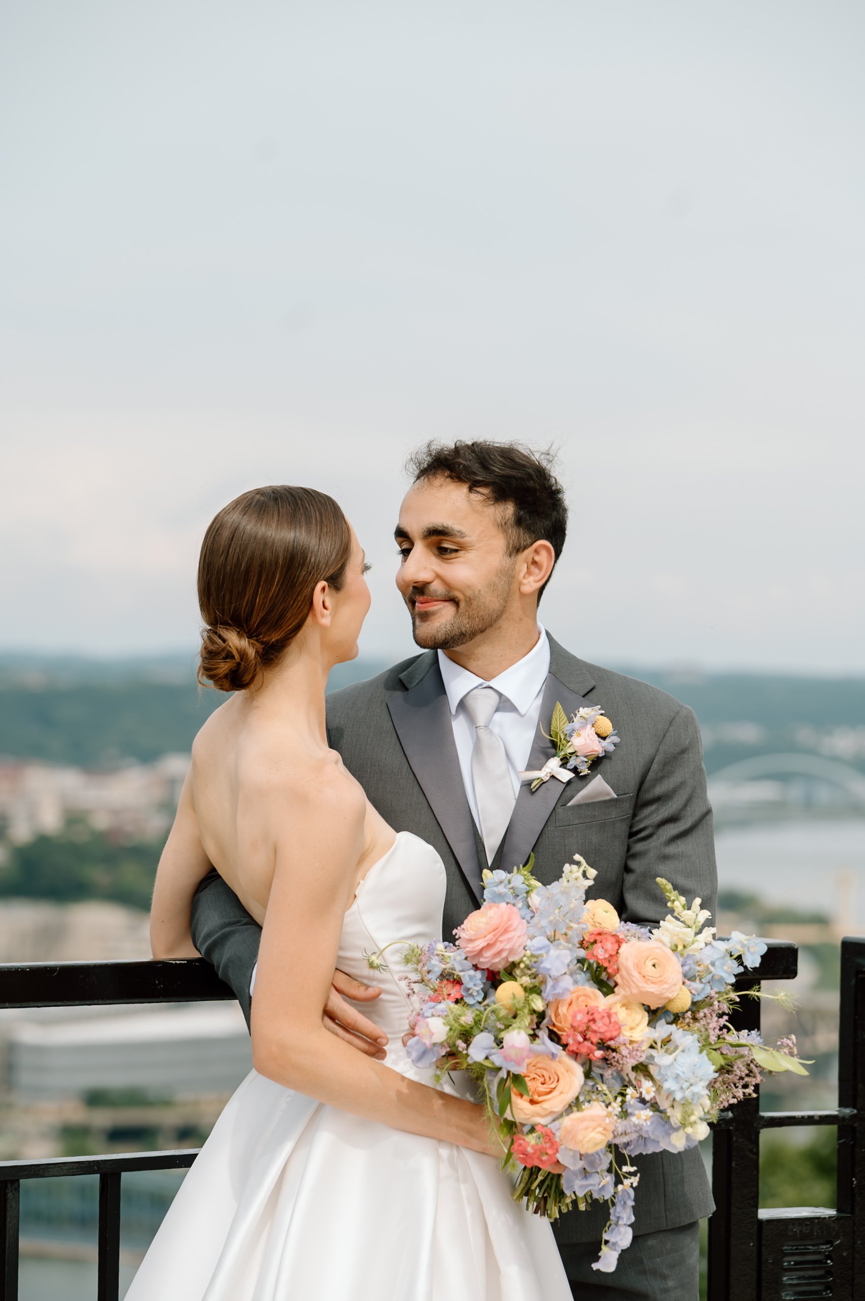 A bride and groom on a balcony, looking at each other. The bride holds a large bouquet of pastel-colored flowers. The groom wears a grey suit, white shirt, and a boutonniere. The background shows a cityscape and water.