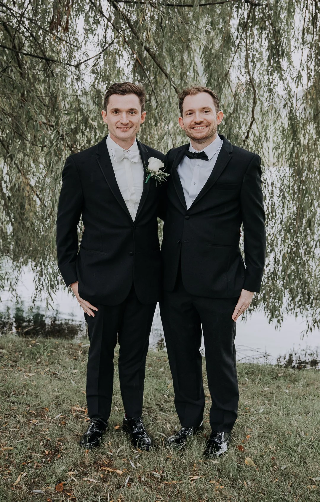 Two men dressed in black tuxedos standing outdoors near a body of water, with trees in the background, smiling at the camera.