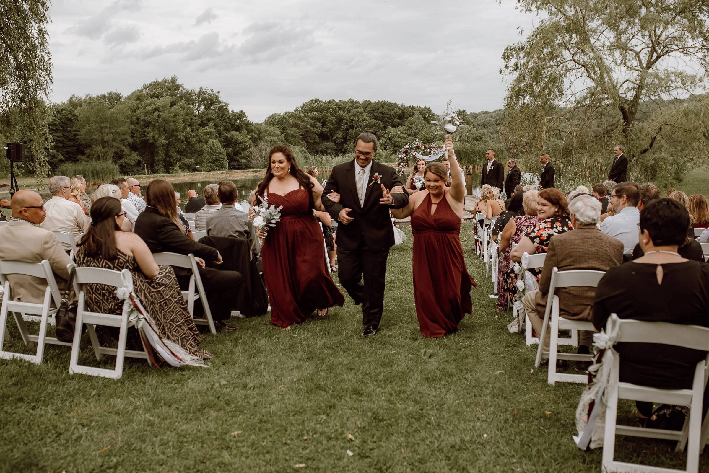 A wedding procession by a lake with guests seated on white chairs watching. The bride and groom walk down the aisle, with the bride in a maroon dress holding a bouquet, and the groom in a black suit. Bridesmaids in matching maroon dresses walk alongs