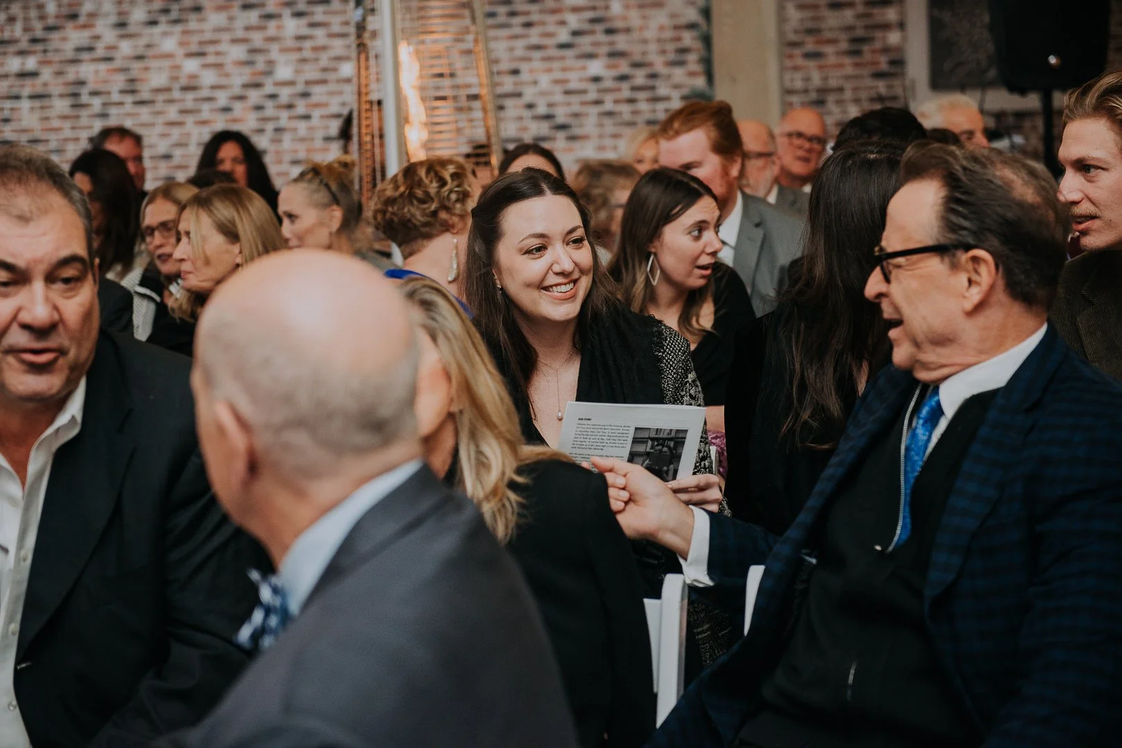 A large group of people at a formal event, talking and interacting, some holding papers, in a venue with brick walls and warm lighting.