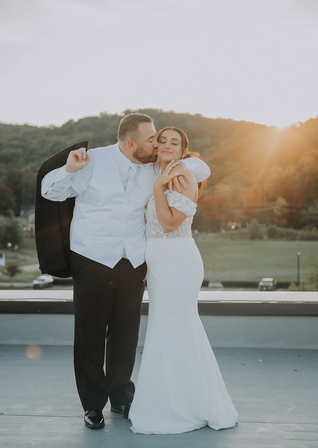 A newlywed couple, the groom in a white shirt and black jacket, and the bride in a white lace wedding gown, sharing a kiss during sunset outdoors.