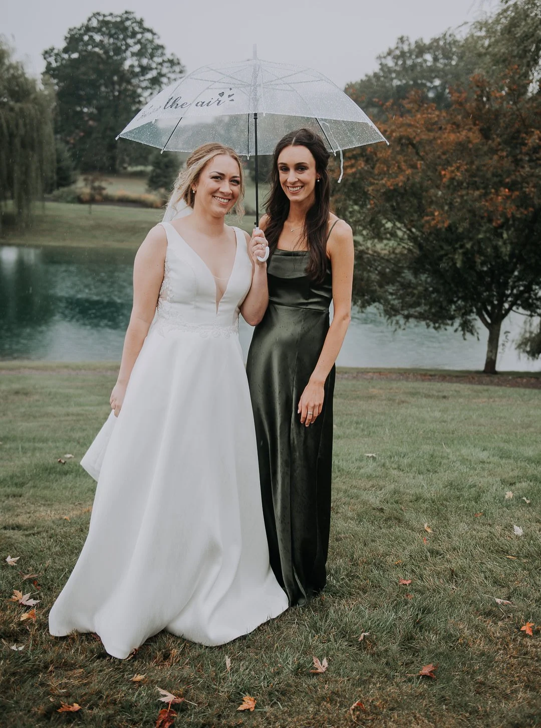 Two women stand outdoors on a rainy day, sharing a clear umbrella. One woman is dressed in a white wedding gown, and the other in a dark satin dress, both smiling.