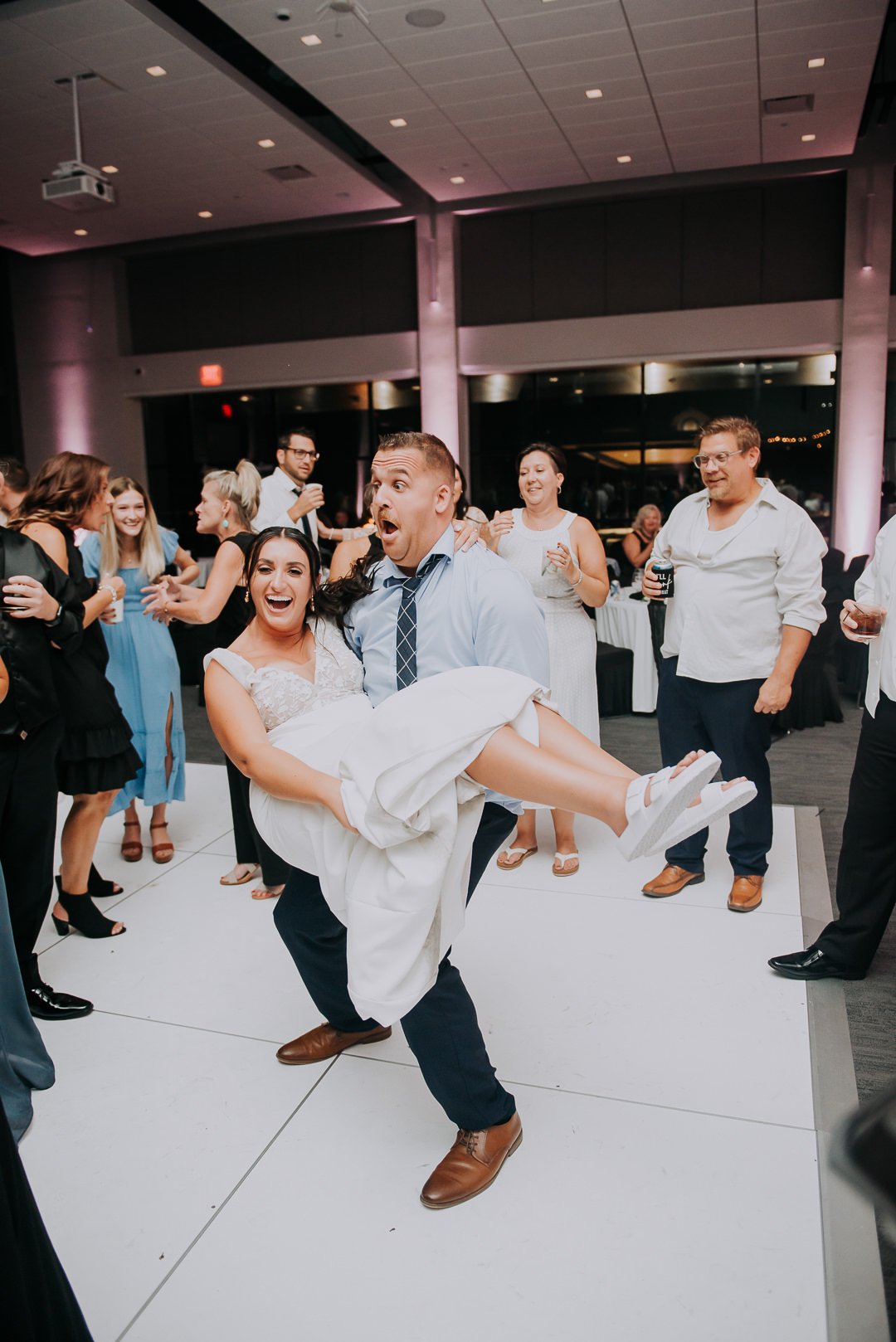 Groom lifting bride during wedding reception dance surrounded by guests enjoying celebration.