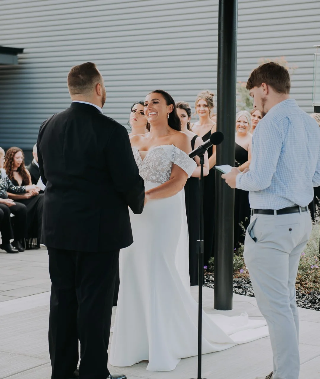A wedding ceremony with a bride and groom exchanging vows, surrounded by witnesses and guests, outdoors near a modern building.