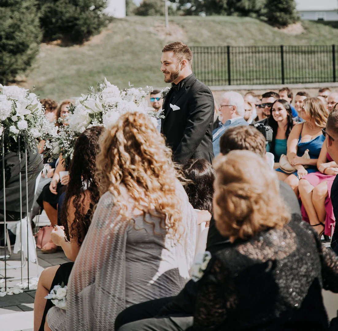 A groom in a black suit with a white pocket square standing at an outdoor wedding ceremony surrounded by seated guests and white floral arrangements.