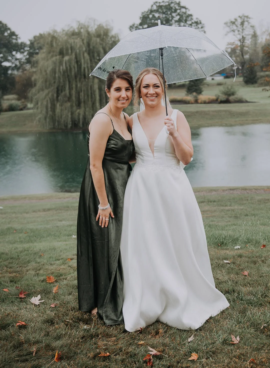 Two women, one in a black dress and one in a white wedding gown, standing on grass near a lake on a rainy day, sharing an umbrella.