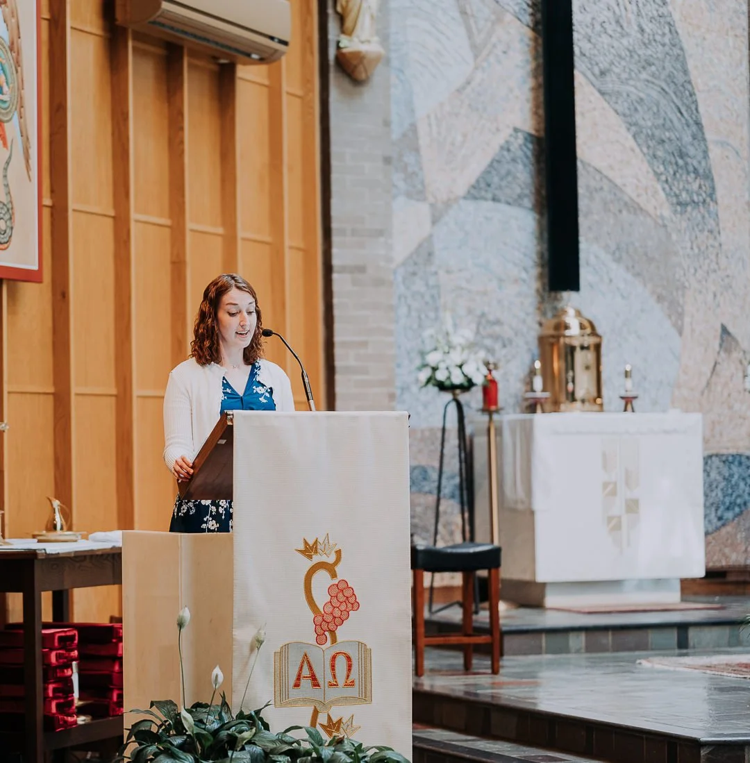 A young woman with curly brown hair stands at a wooden lectern decorated with the Greek letters Alpha and Omega, speaking into a microphone during a church service inside a church with wooden paneling and a mosaic wall. The altar with candles and a f